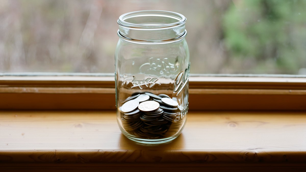 A glass jar half filled with loose coins standing on a windowsill